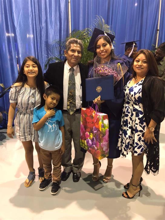 A proud graduate stands with family members while holding a diploma and flowers.