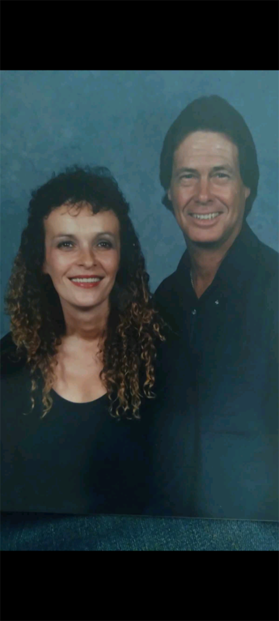 A happy couple stands close together, both wearing dark clothing and smiling warmly.