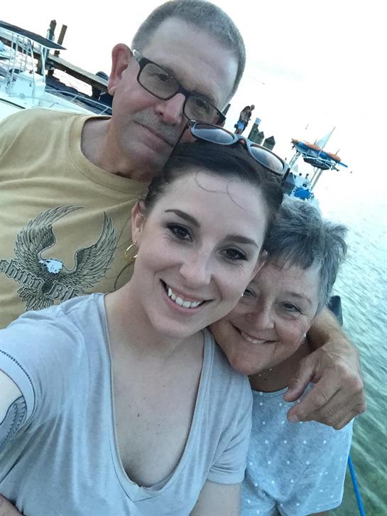 A young woman poses with her grandparents, smiling happily on a boat at dusk.