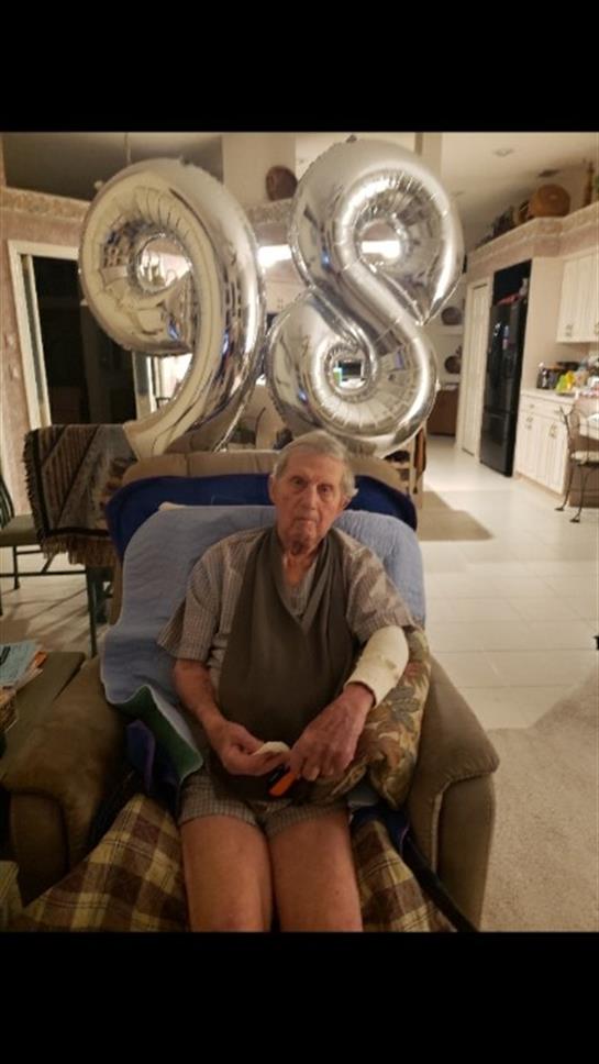 An elderly man celebrates his 98th birthday in a chair with balloons behind him.