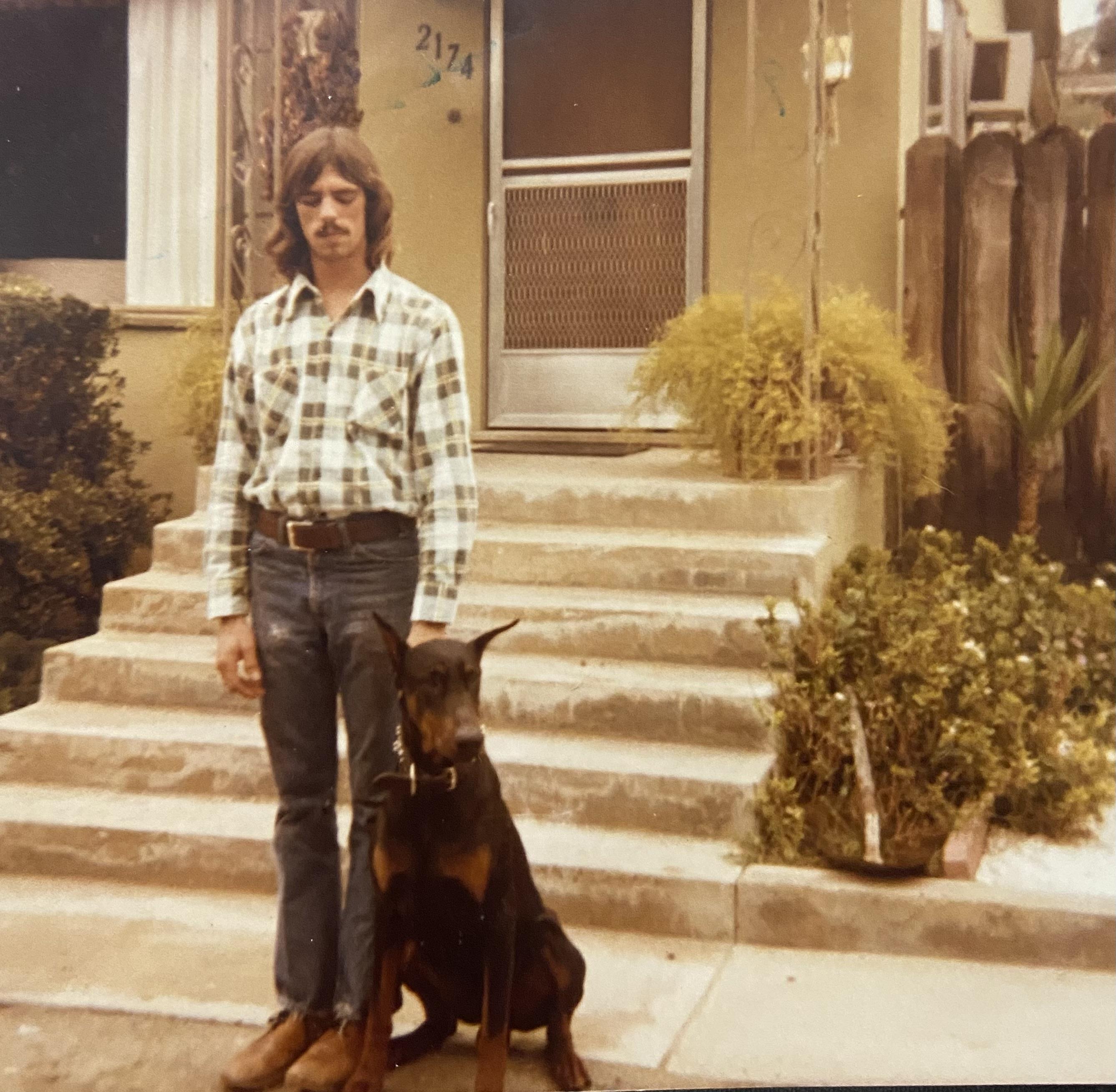 A man stands next to a dog on steps outside a house in a residential area during the day.