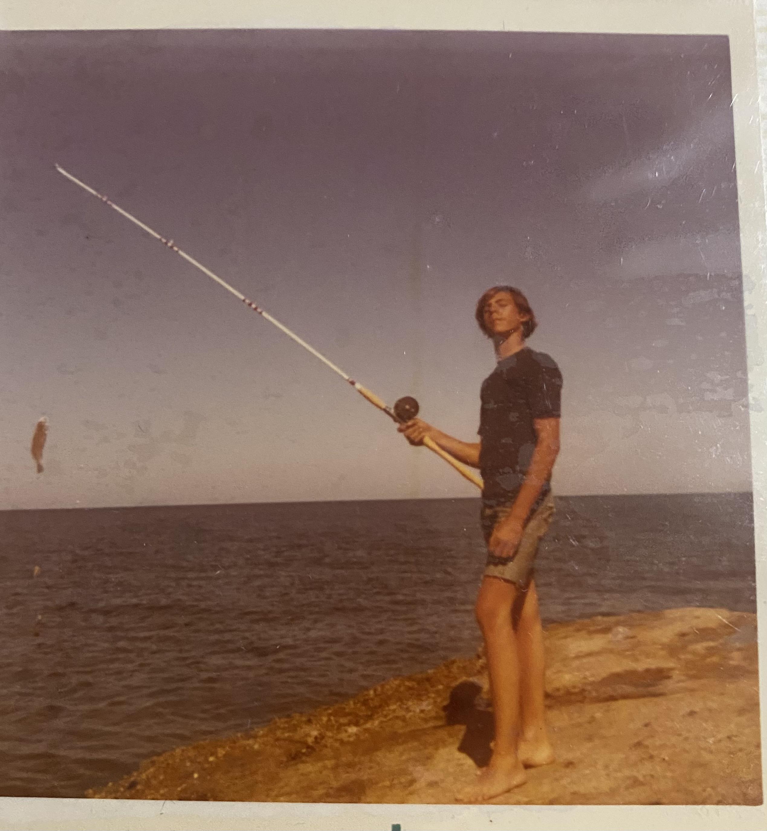 A man stands on the shore with a fishing rod, looking out over the water with a fish on the line.