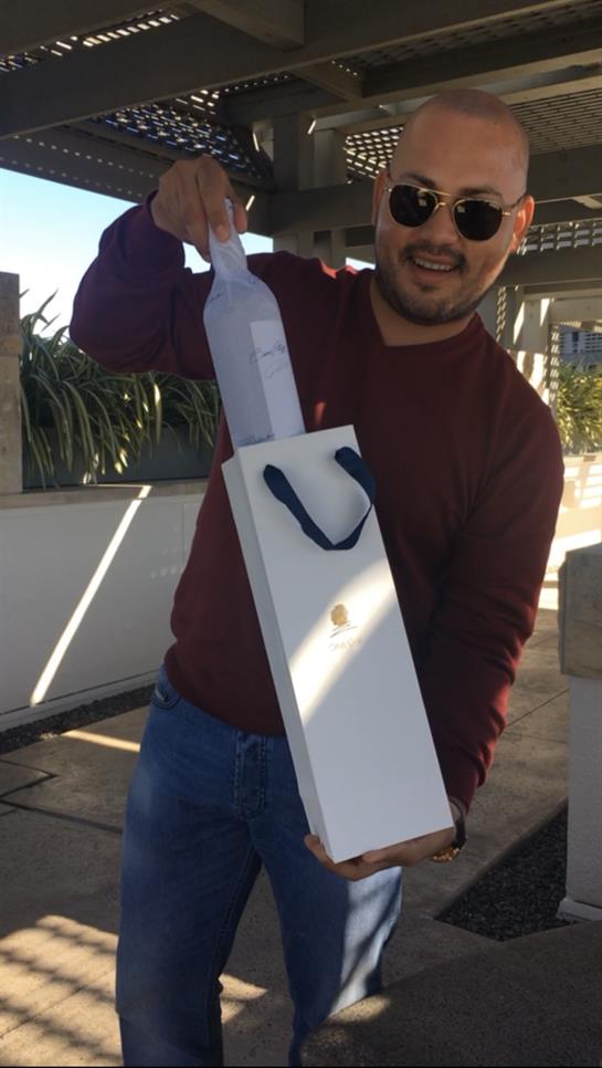 A man stands outside, cheerfully presenting a gift-wrapped bottle, enjoying the sunny weather.