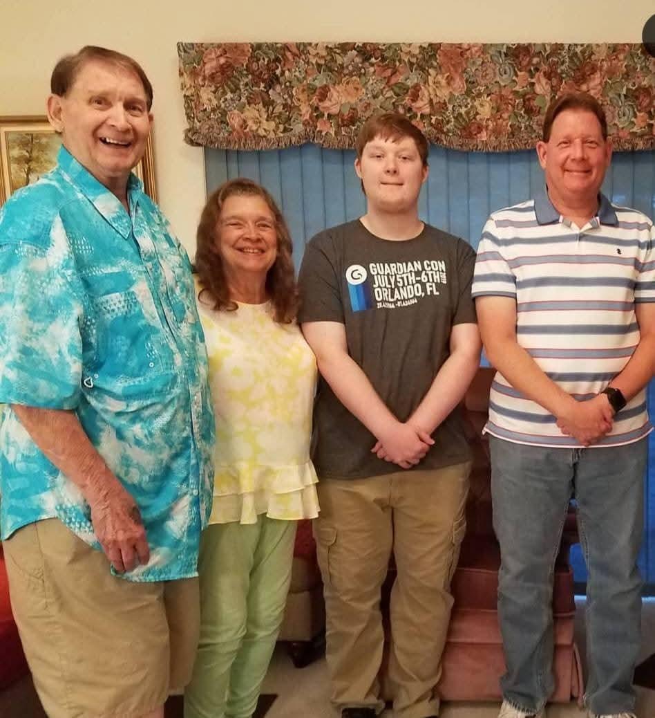 Four family members are gathered indoors, smiling together in casual clothing on a summer day.