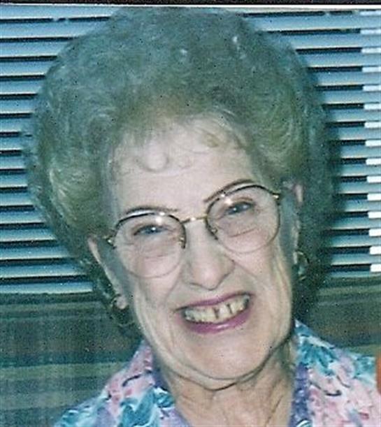 Elderly woman with glasses beams joyfully while seated at home, wearing colorful attire.