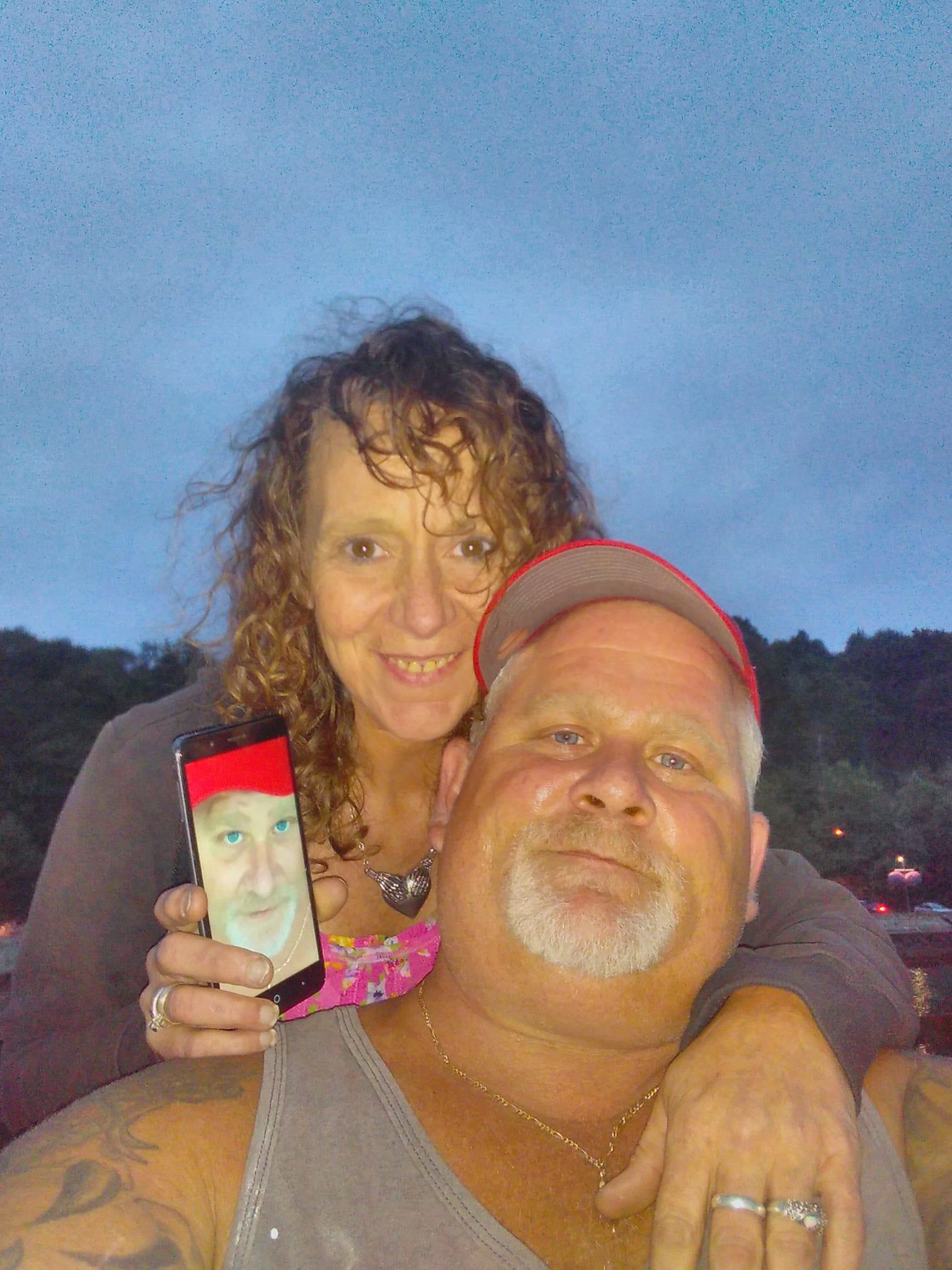 A couple smiles together, one holding a phone displaying a funny face, during sunset.