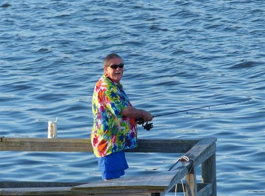 A man in a colorful shirt enjoys fishing from a wooden dock on a sunny day.