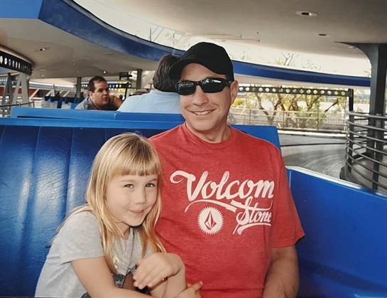 A cheerful father and daughter sit together on a ride, smiling and enjoying their day.