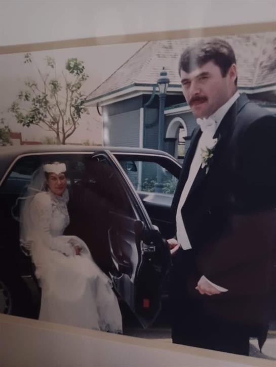 Bride in white dress and groom in suit interact near vehicle at wedding venue.