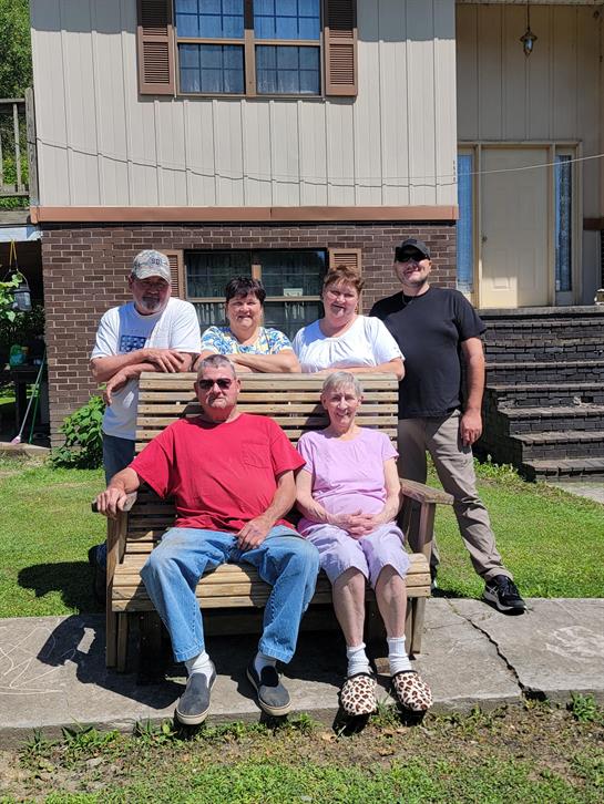 Six family members pose together, smiling on a rustic bench outside their house in bright sunlight.