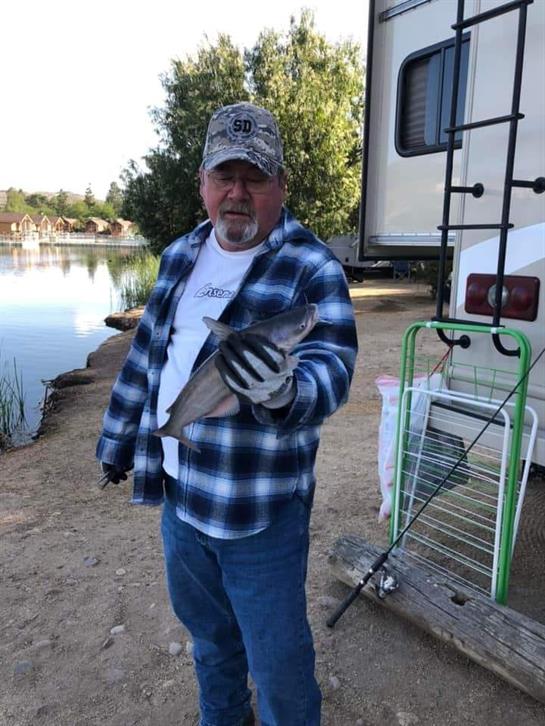 A man stands near a lake, proudly displaying a fish he just caught while camping.