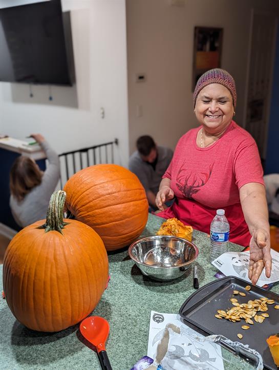 A woman prepares pumpkin seeds while family members engage in carving pumpkins.