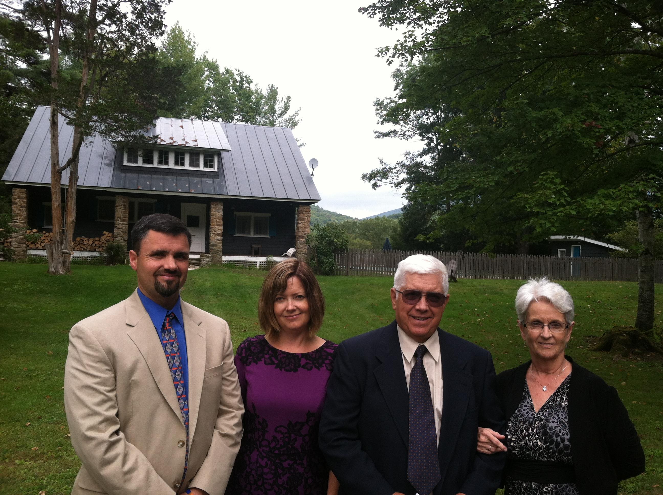 Four family members pose together outside a rustic house on a cloudy day in nature's tranquility.