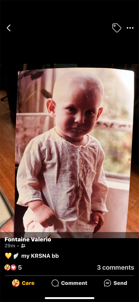A toddler with a bald head stands by a window, wearing a light shirt and gazing thoughtfully.