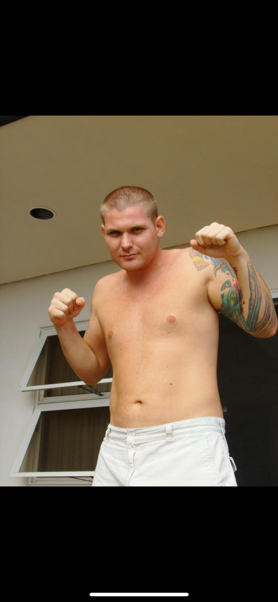 A man displays a fighting stance outside a contemporary structure, showcasing a tattoo.