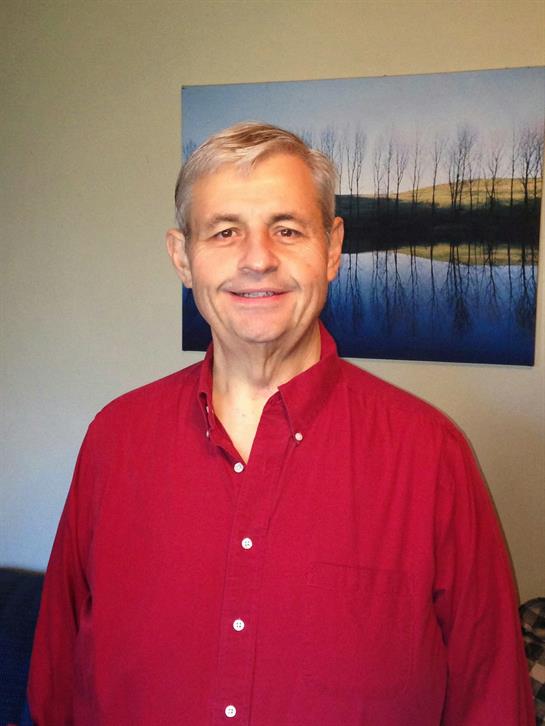 A man with gray hair is smiling in a red shirt, standing in a room with lake art.