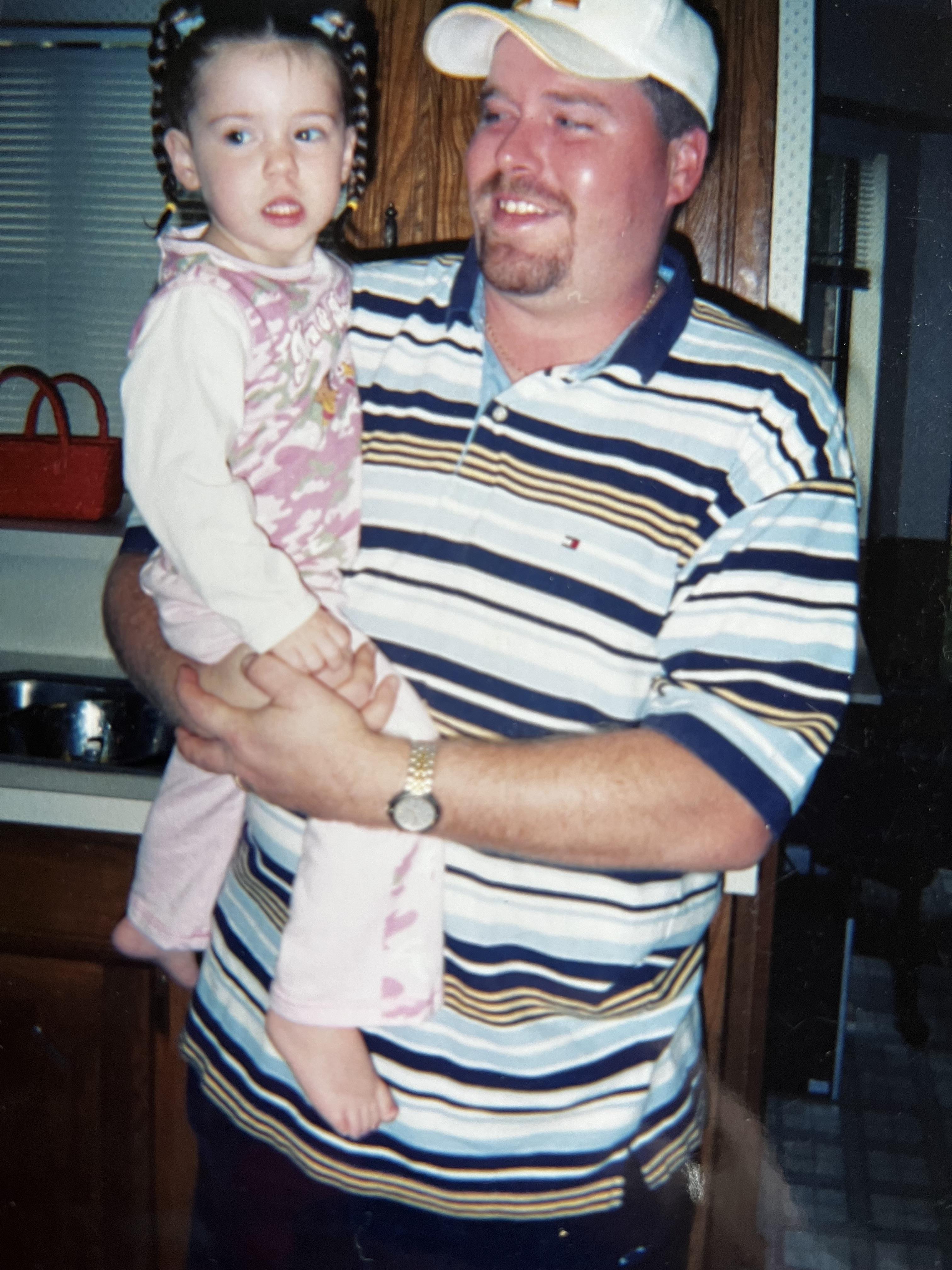 A man holds his young daughter inside a cozy kitchen, both smiling and enjoying their time together.
