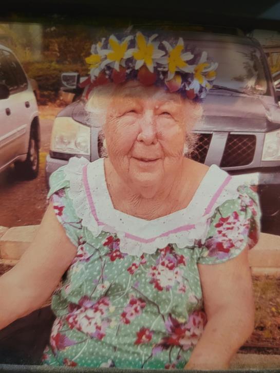 A joyful elderly woman in floral attire sits in a garden, wearing a flower crown and smiling.