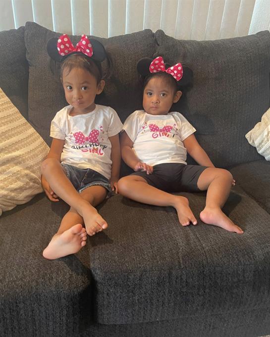 Two young girls dressed in identical outfits sit comfortably on a gray couch.