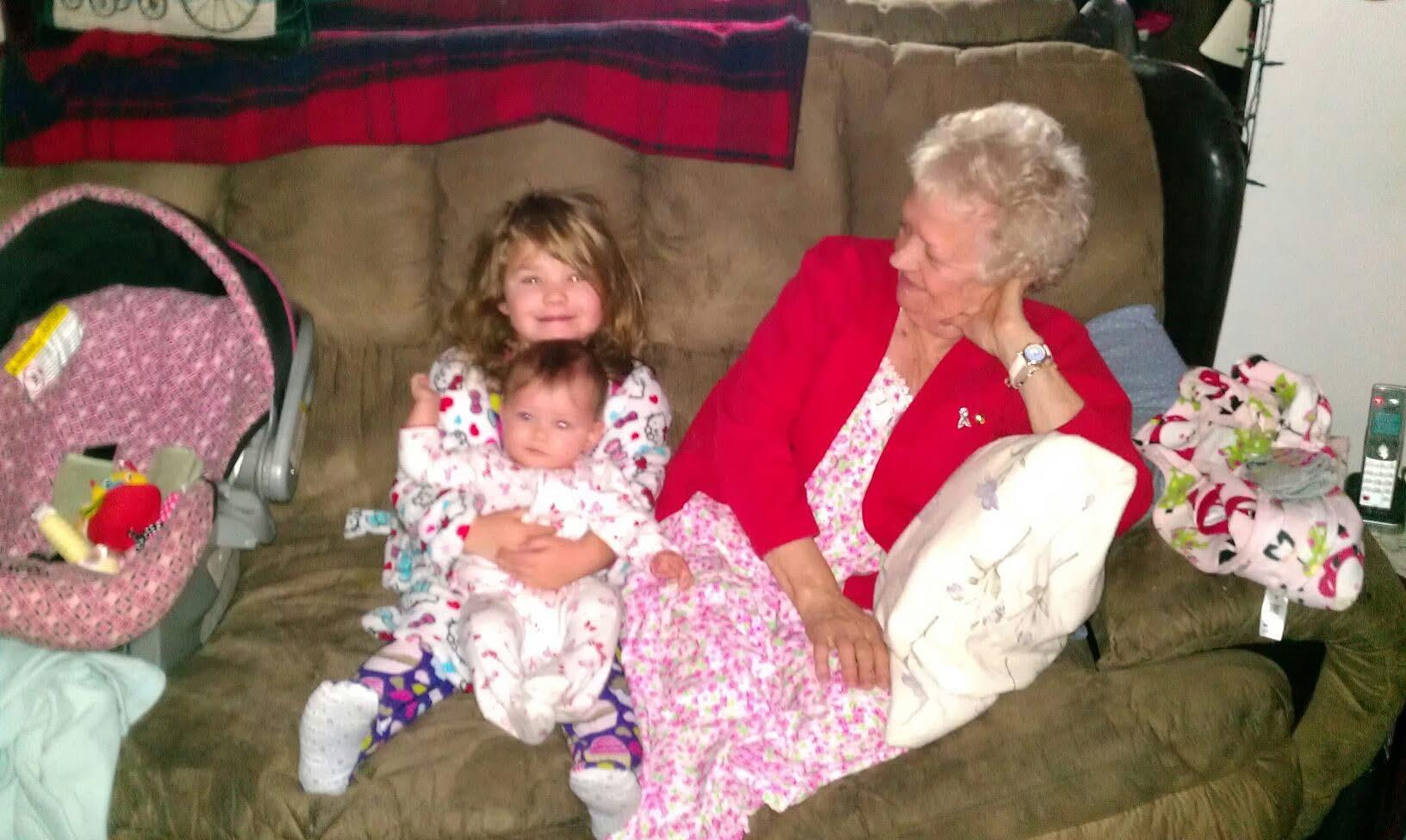 Grandmother sits comfortably on a couch, embracing her grandchildren in colorful pajamas.
