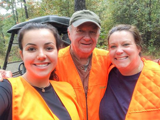 Three individuals smile while wearing bright orange safety vests in a forest setting.