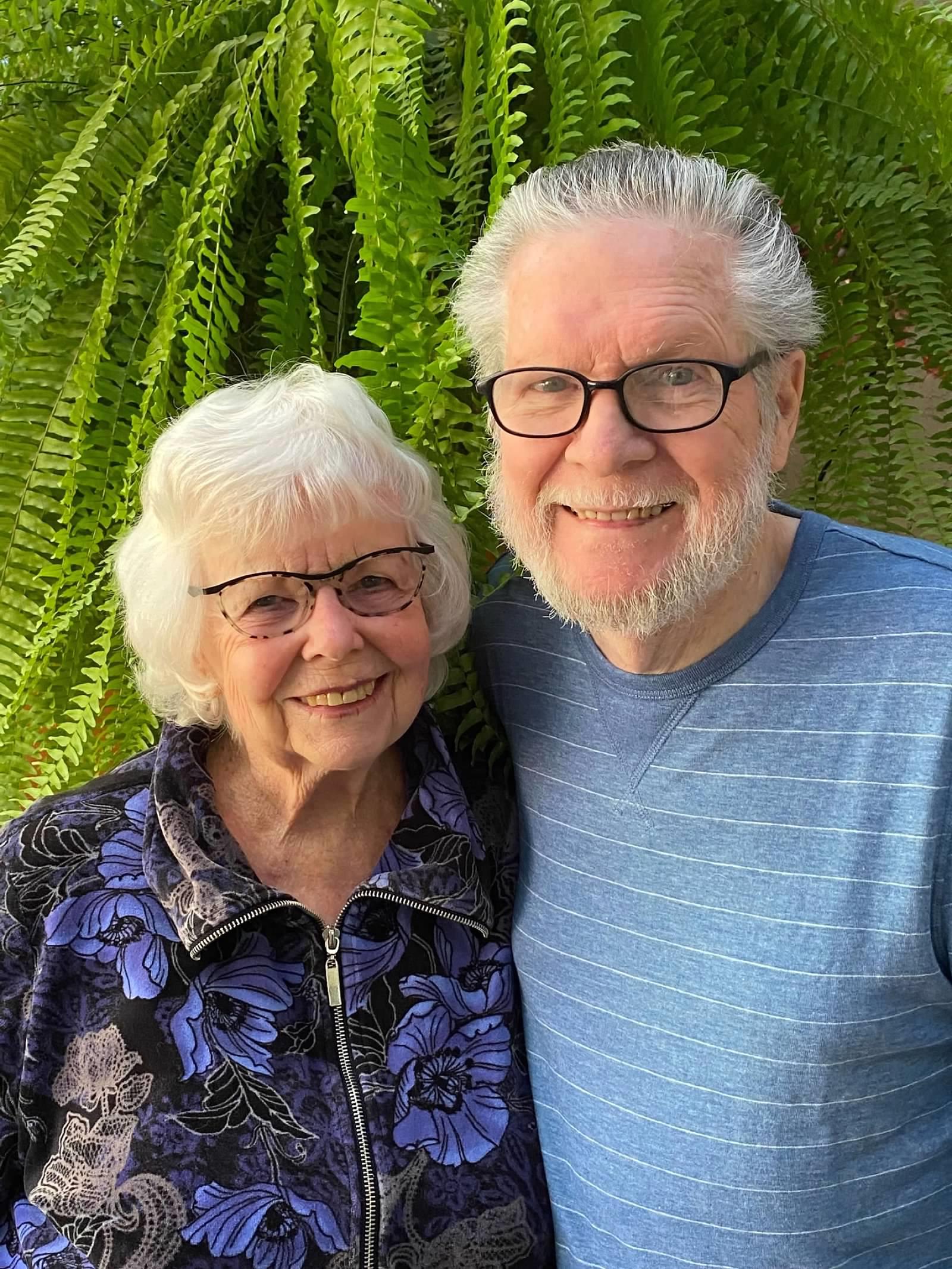Two seniors pose cheerfully together with vibrant ferns in the background on a sunny day.