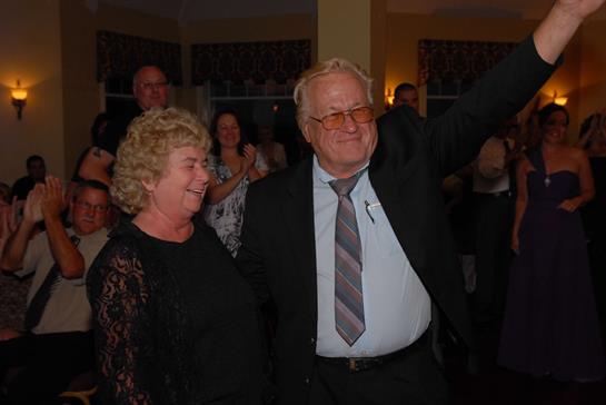 An elderly couple enjoys a joyful moment at a celebration, surrounded by smiling friends and family.
