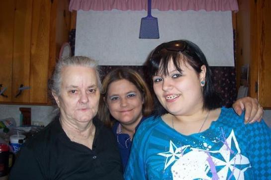 Three women of different generations stand together, all smiling, in a warm kitchen setting.