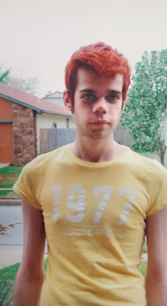 Outside a suburban house, a young man with red hair poses in a yellow shirt showcasing a number.