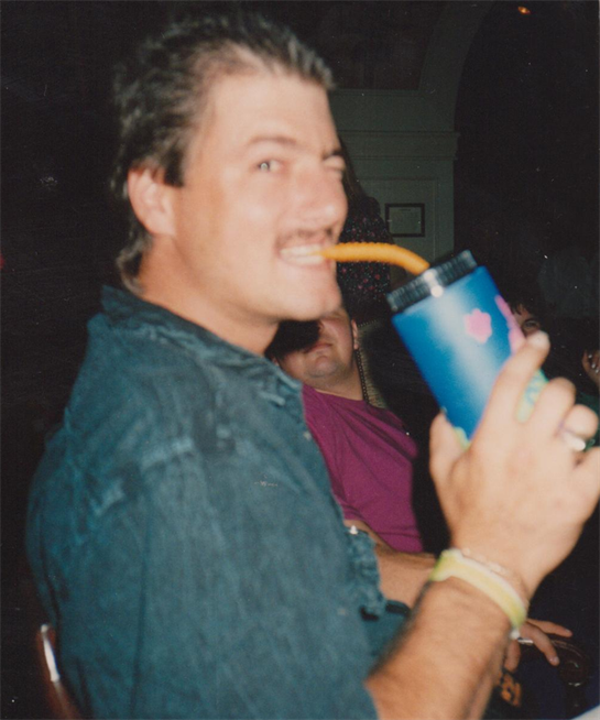 A man smiles while holding a large drink, possibly at a party with friends nearby.