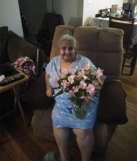 An elderly woman sits comfortably in a recliner, displaying joy with pink flowers in hand.