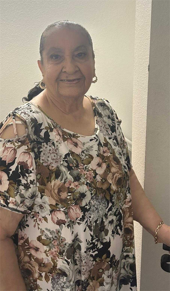 An elder woman with a warm smile poses by her home's doorway, dressed in a floral top.