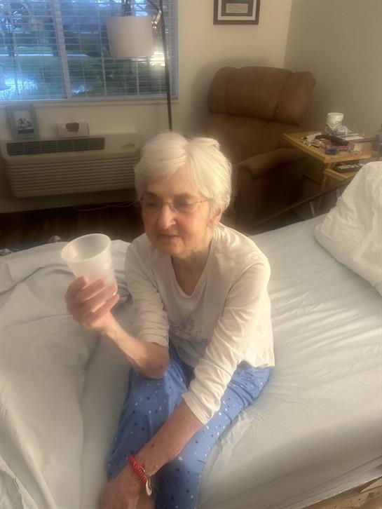 An elderly woman sits on a bed, holding a plastic cup and looking thoughtfully while relaxing.