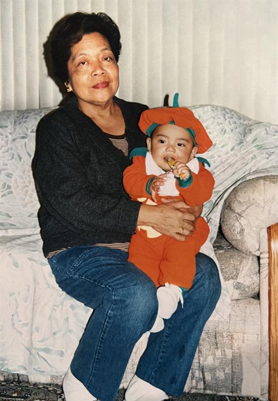 An elderly woman smiles warmly while holding a cheerful toddler in a playful outfit by the couch.