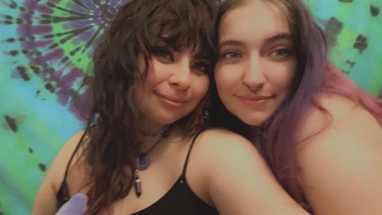 Two young women smile together in a playful pose against a vibrant backdrop.