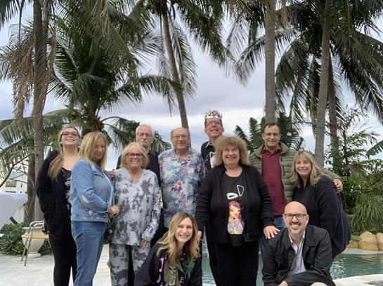 Friends gather joyfully in a tropical setting surrounded by palm trees for a celebration.