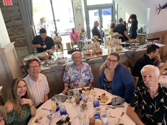 A group of friends smiles around a table filled with diverse brunch dishes and drinks.