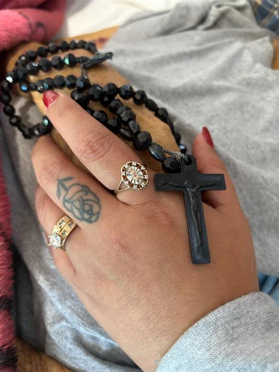 A hand with rings and tattoos holds black prayer beads with a cross pendant.