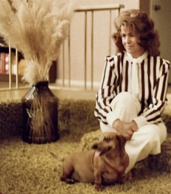 Woman interacts with small brown dog while sitting on a carpet near a decorative plant.