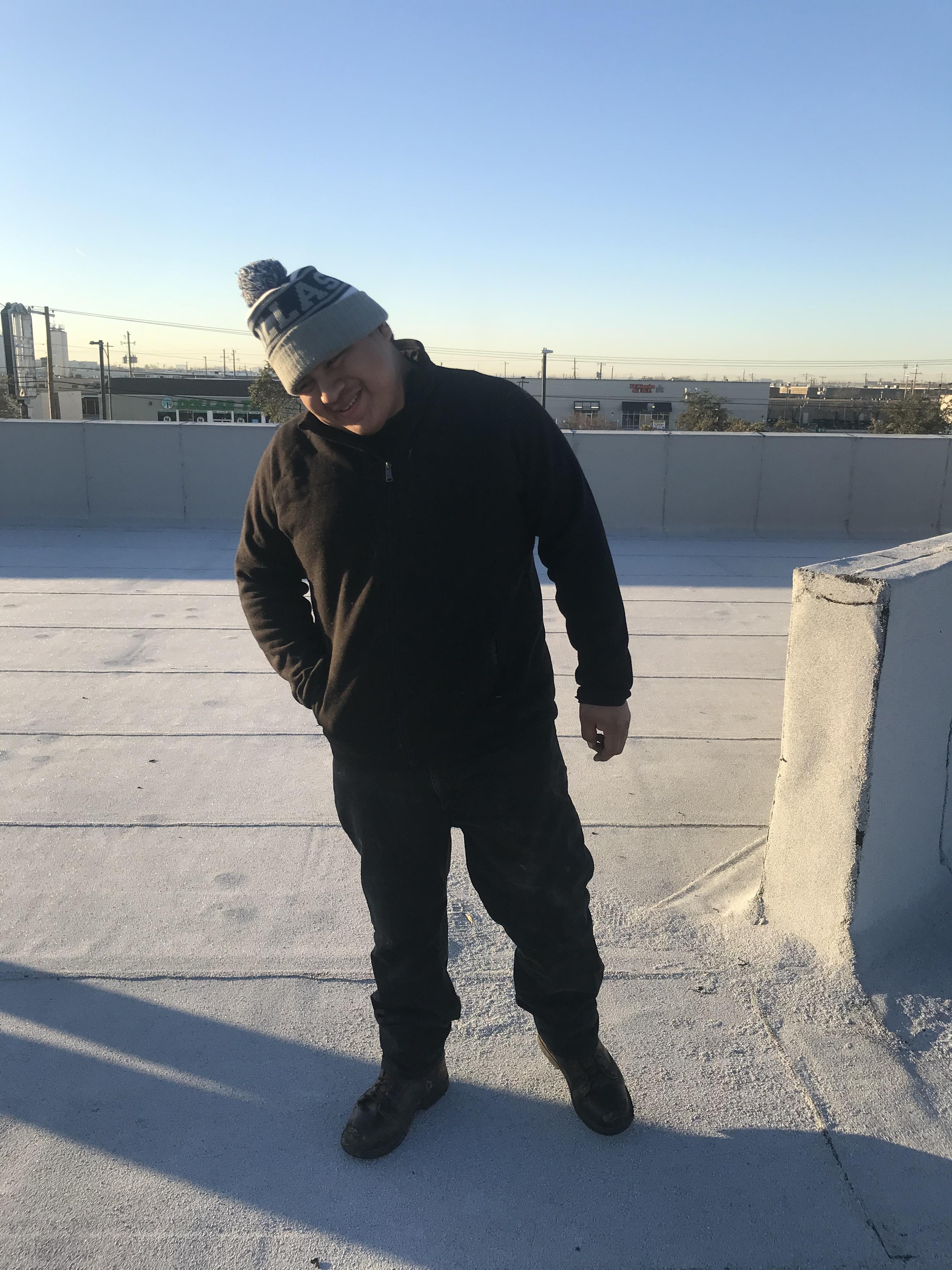 Individual dressed in black stands on a rooftop under a sunny sky, showcasing a relaxed posture.