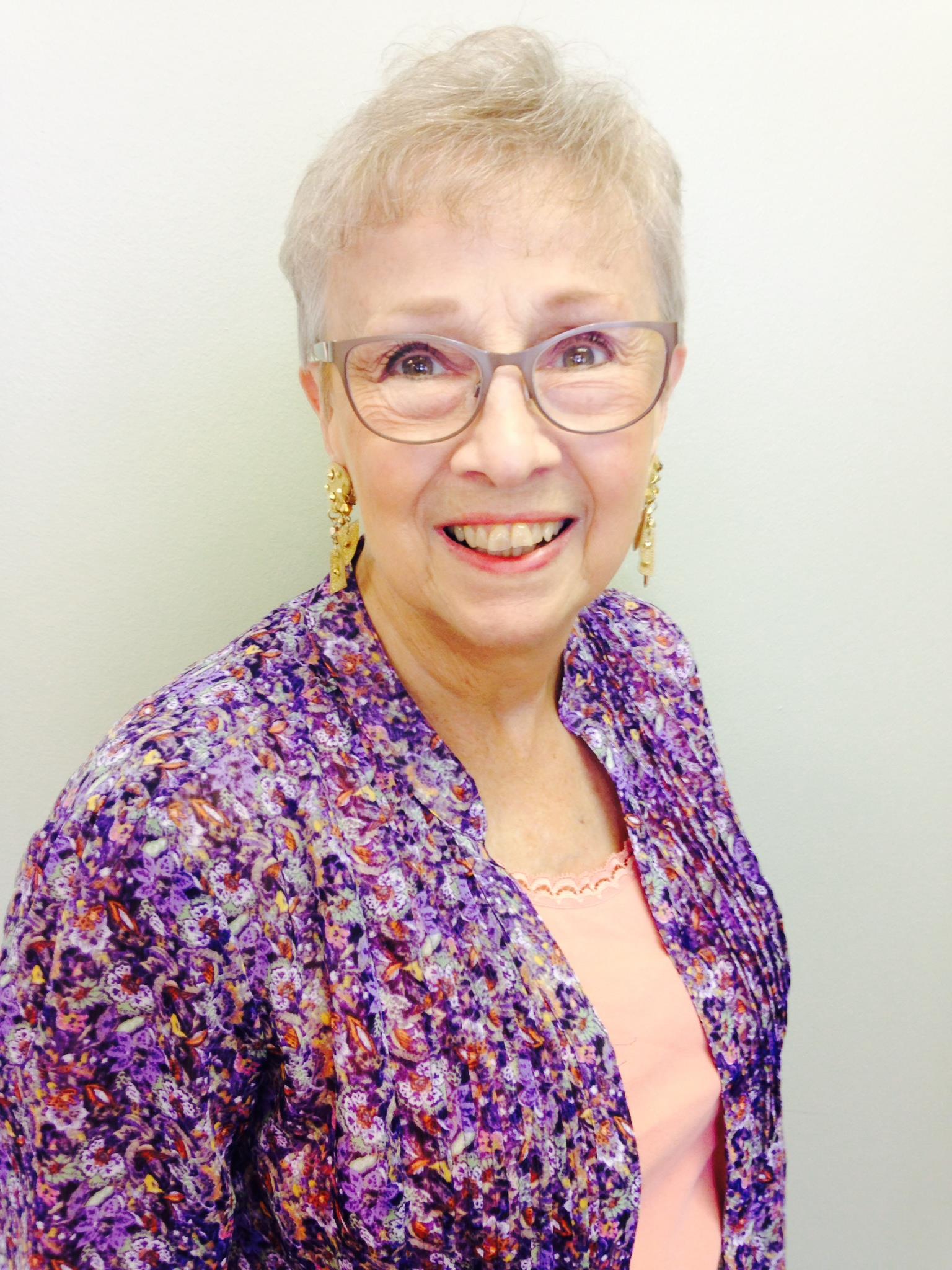Elderly woman with short gray hair and glasses poses happily, wearing a vibrant purple jacket.