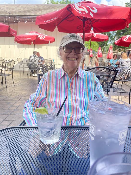 An elderly woman sits at a table enjoying a drink while smiling in a lively cafe atmosphere.