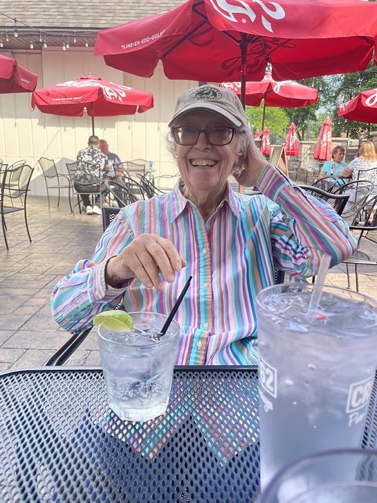 A cheerful elderly man is savoring a cold drink while seated outdoors, surrounded by umbrellas.