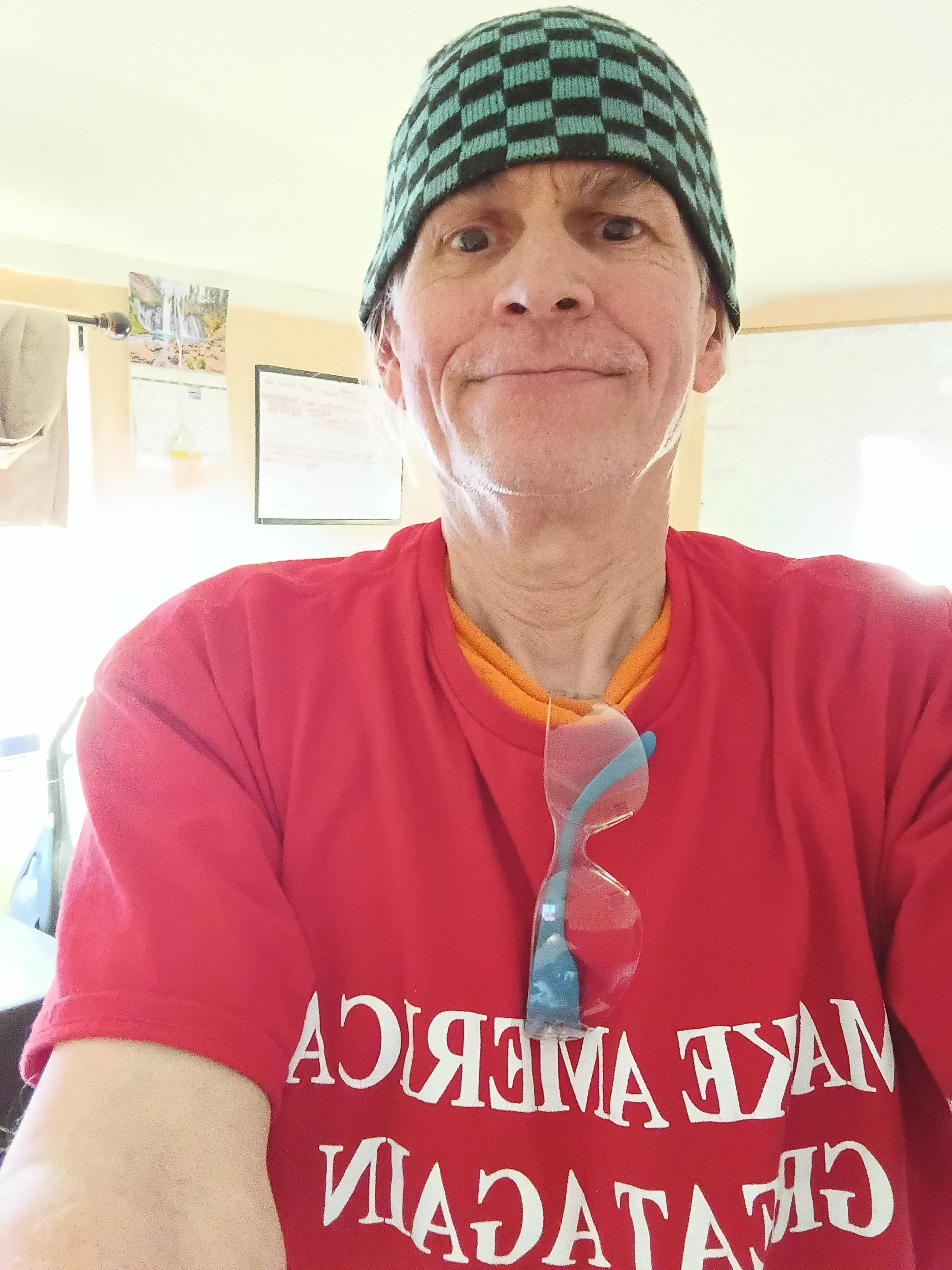 Individual in a red shirt and checkered hat shows a cheerful expression inside a well-lit room.