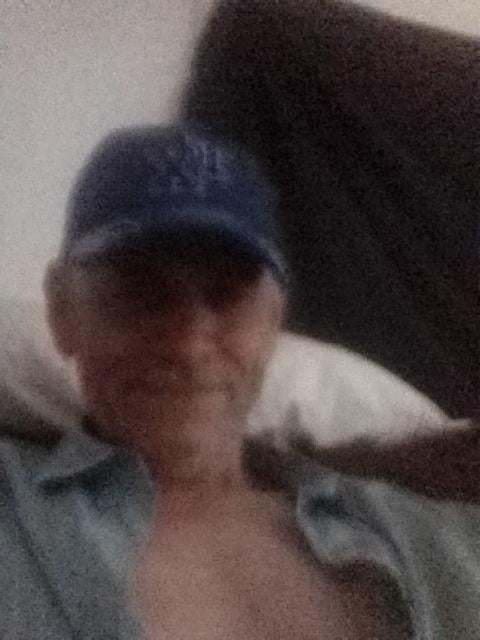 An older man with a blue cap enjoys a relaxed moment on a sofa indoors, showing a warm smile.