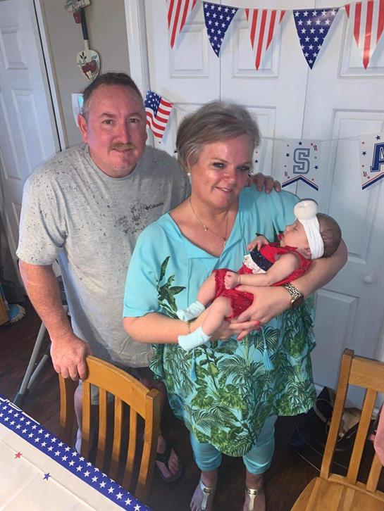 A couple joyfully holds their newborn baby, dressed in a festive outfit, at a home celebration.