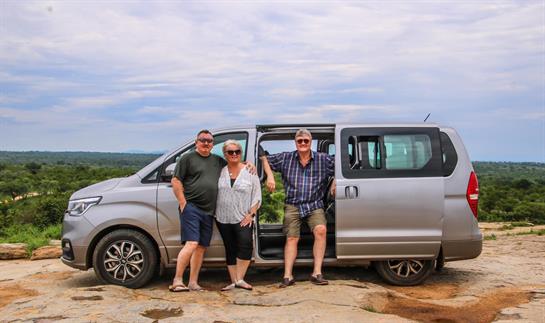 Group of three travelers posed beside a silver van, taking in the beautiful landscape.