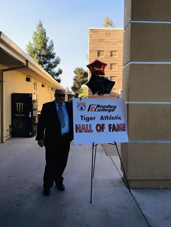 A man in a suit stands beside a sign for the Tiger Athletic Hall of Fame at Reedley College.