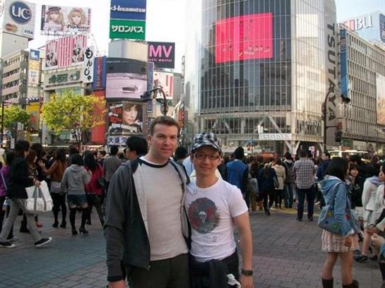 Two friends pose in front of lively Shibuya Crossing surrounded by a large crowd.