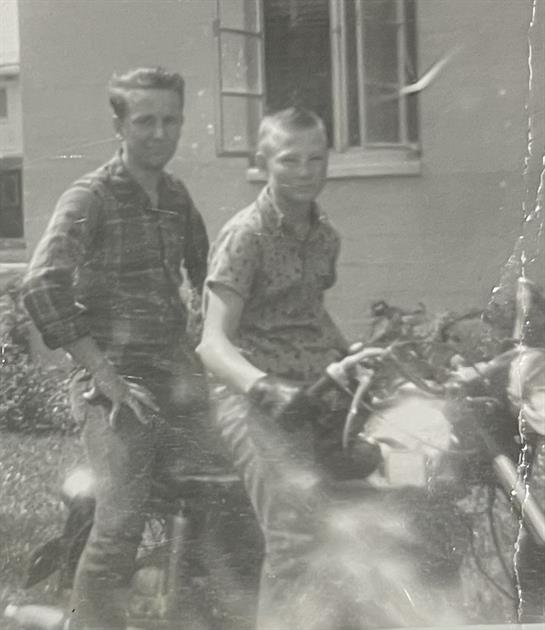 Two boys sit on a motorcycle, laughing and smiling under a warm sun near a home.
